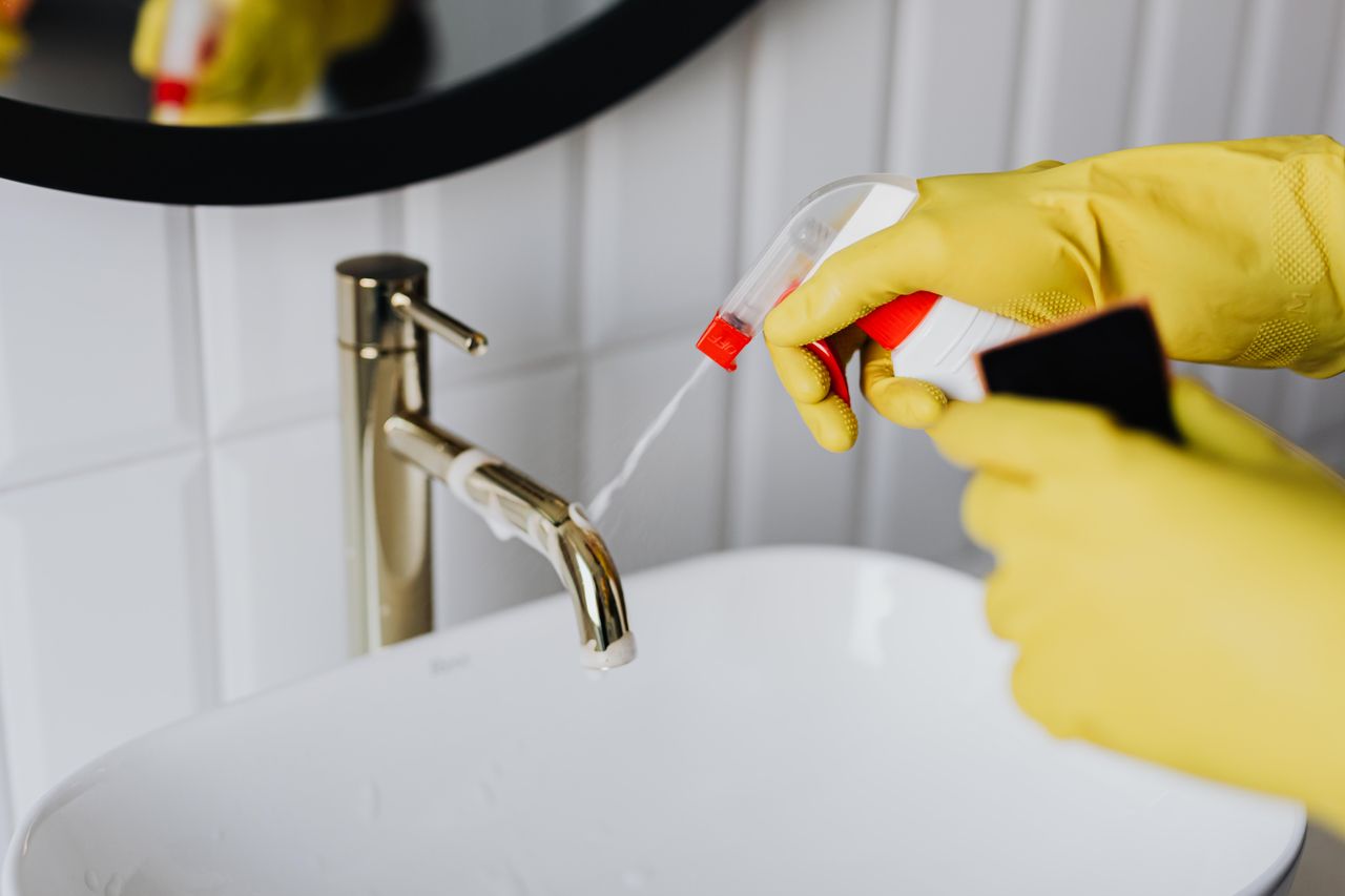 services-02 From above anonymous housekeeper in yellow rubber gloves spraying cleaner to chrome water faucet in bathroom