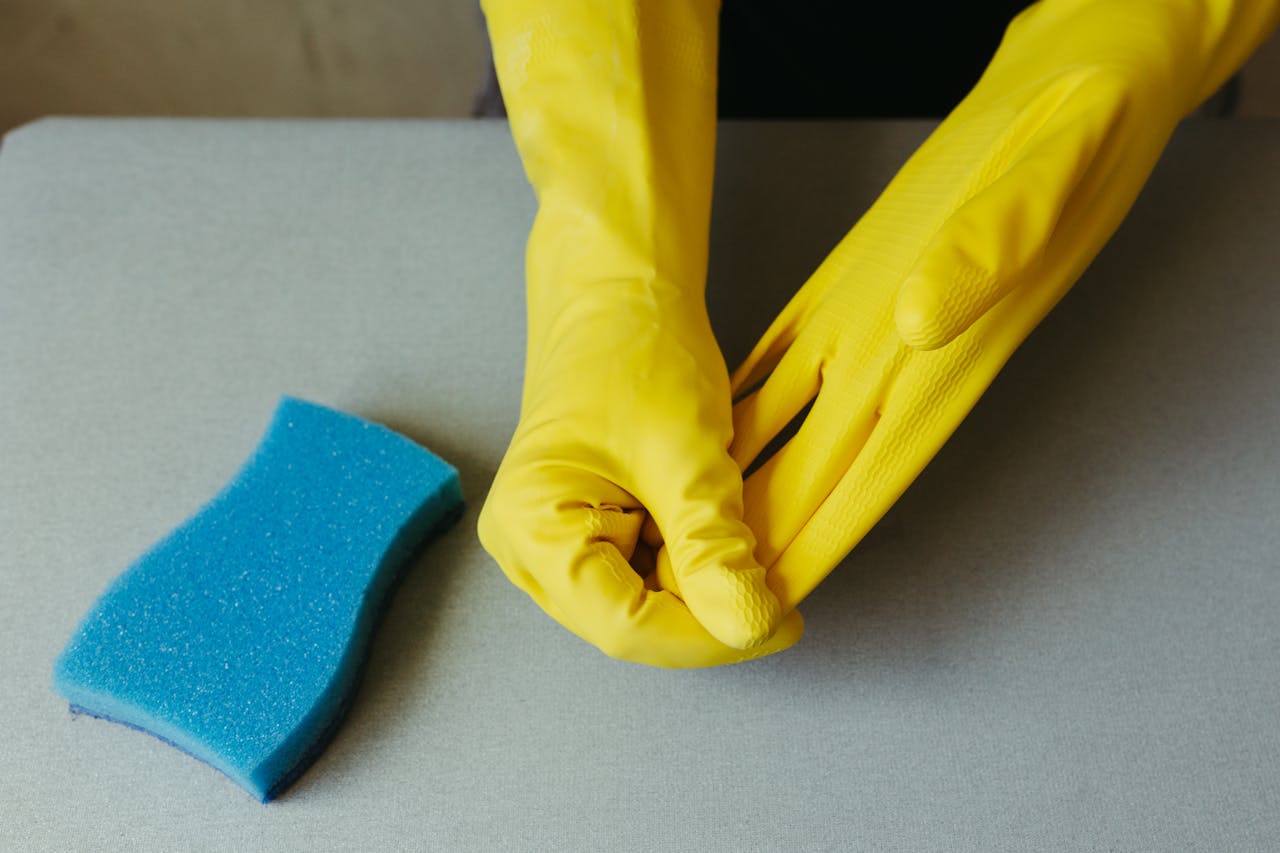 about-01 Close-up of hands wearing yellow gloves next to blue sponge on gray surface.